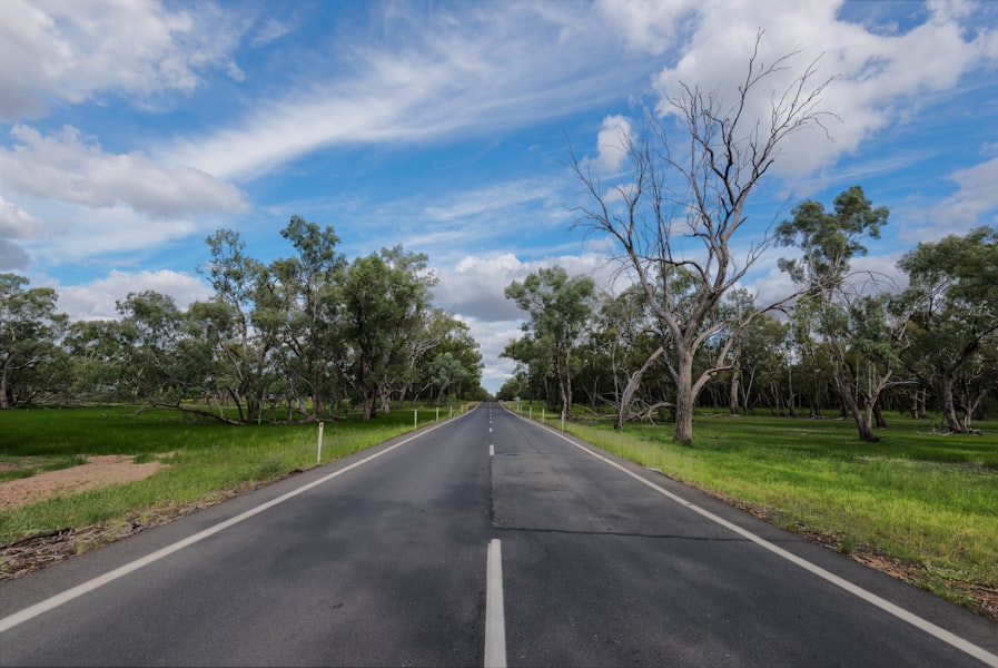 gray asphalt road between green grass field under blue and white sunny cloudy sky during daytime