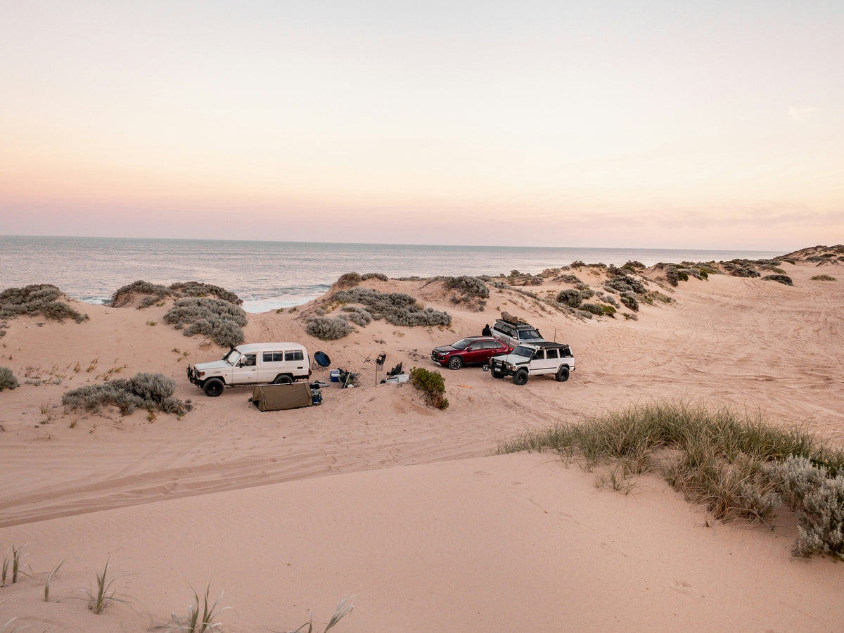 4WDs camped in coastal sand dunes at dusk: a swag, camp chairs and an esky set up beside a LandCruiser